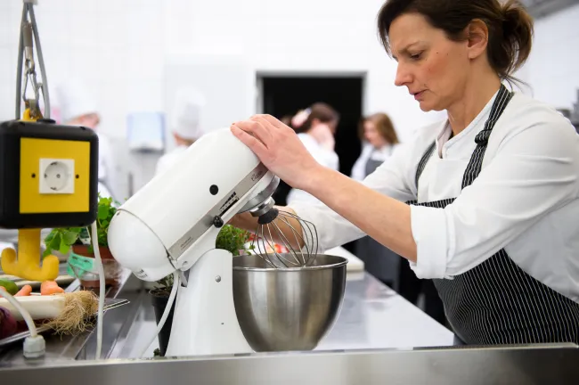 Frau in Kochkleidung nutzt Küchenmaschine zum Rühren in Edelstahlbehälter in moderner Großküche mit Kollegen im Hintergrund​​​​​​​​​​​​​​​​​​​​​​